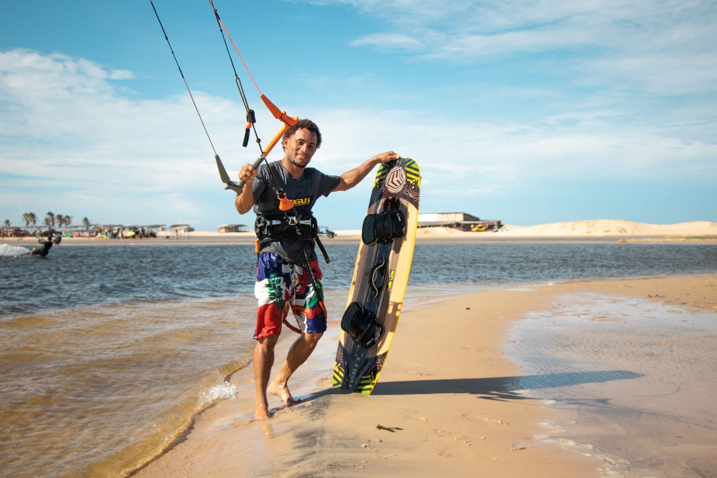 Homem segura uma prancha de kitesurf em uma praia ensolarada à beira-mar