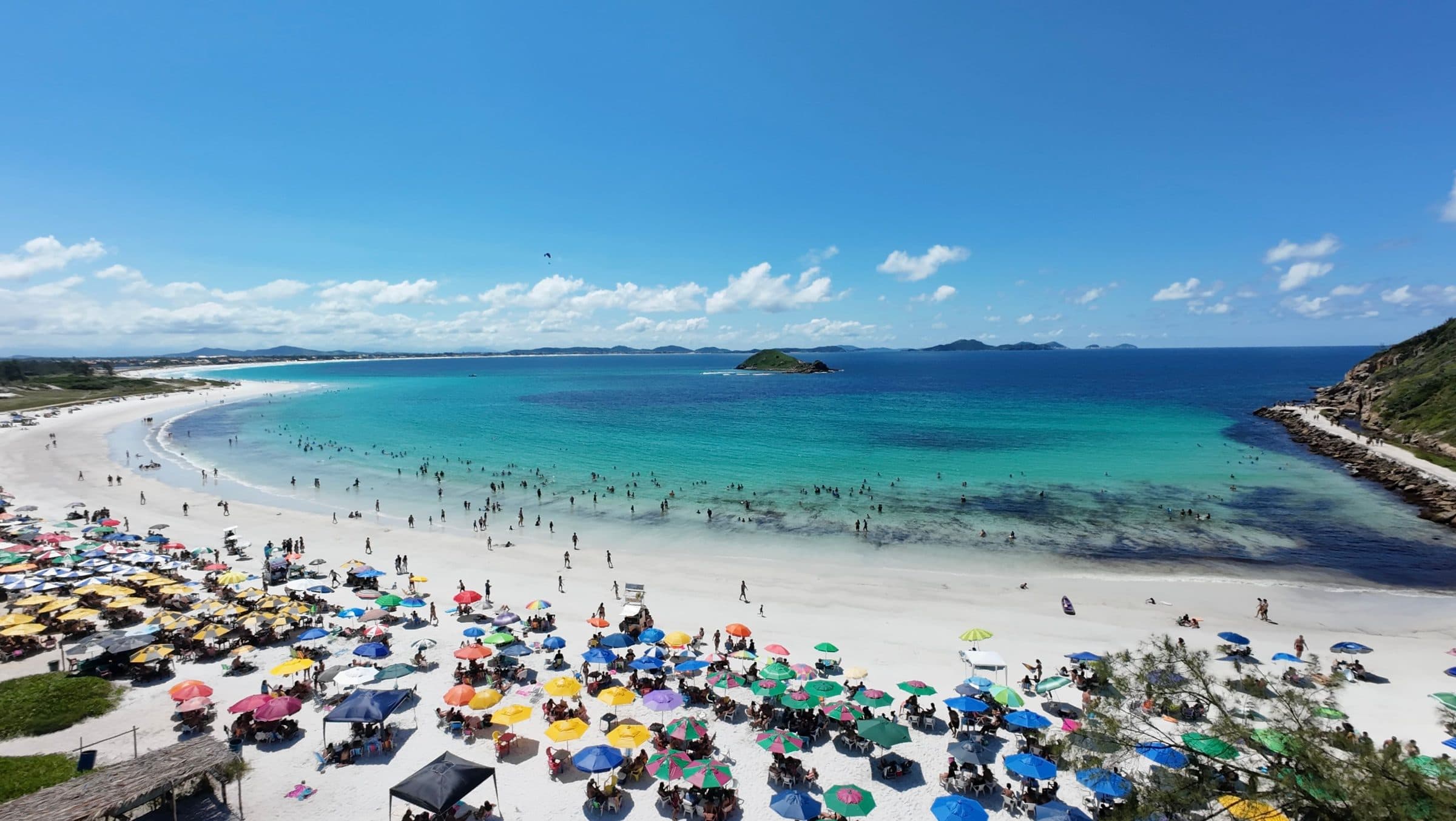 Vista aérea de praia com pessoas e guarda-sóis coloridos espalhados pela areia em Arraial do Cabo
