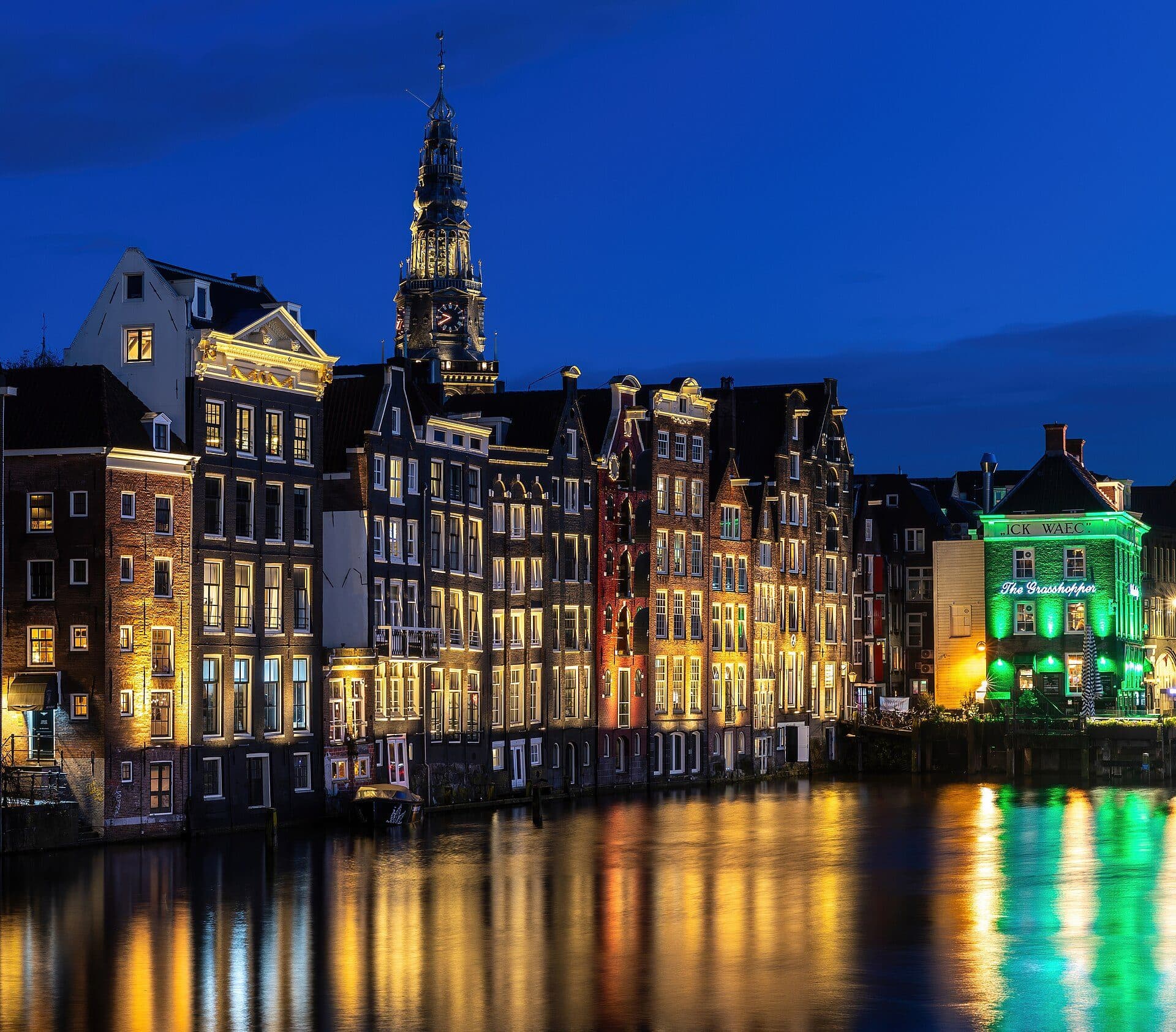Casas de canal e a igreja Oude Kerk refletidas nas águas de Damrak durante a hora azul em Amsterdã