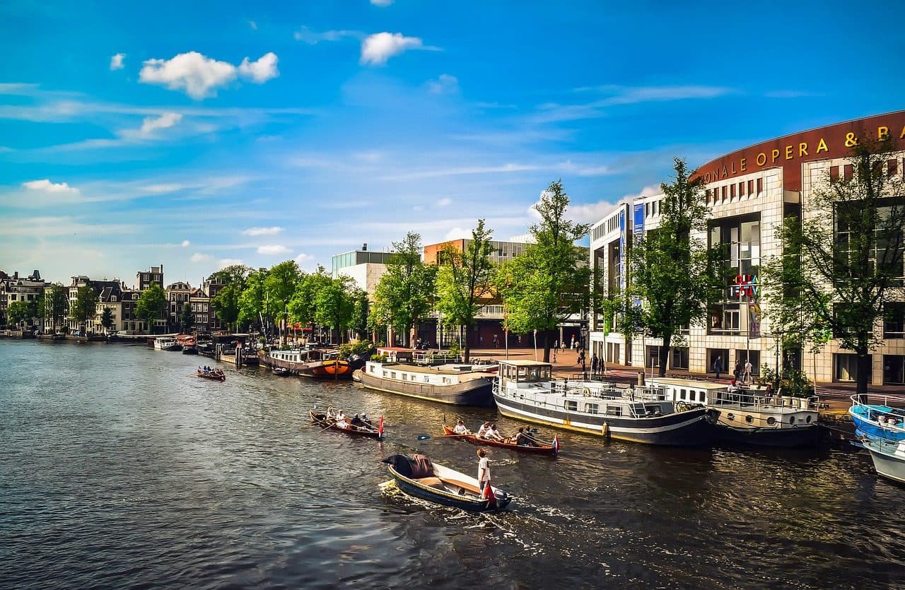 Canal urbano em Amsterdã com barcos na água e prédios ao longo da margem sob céu com nuvens