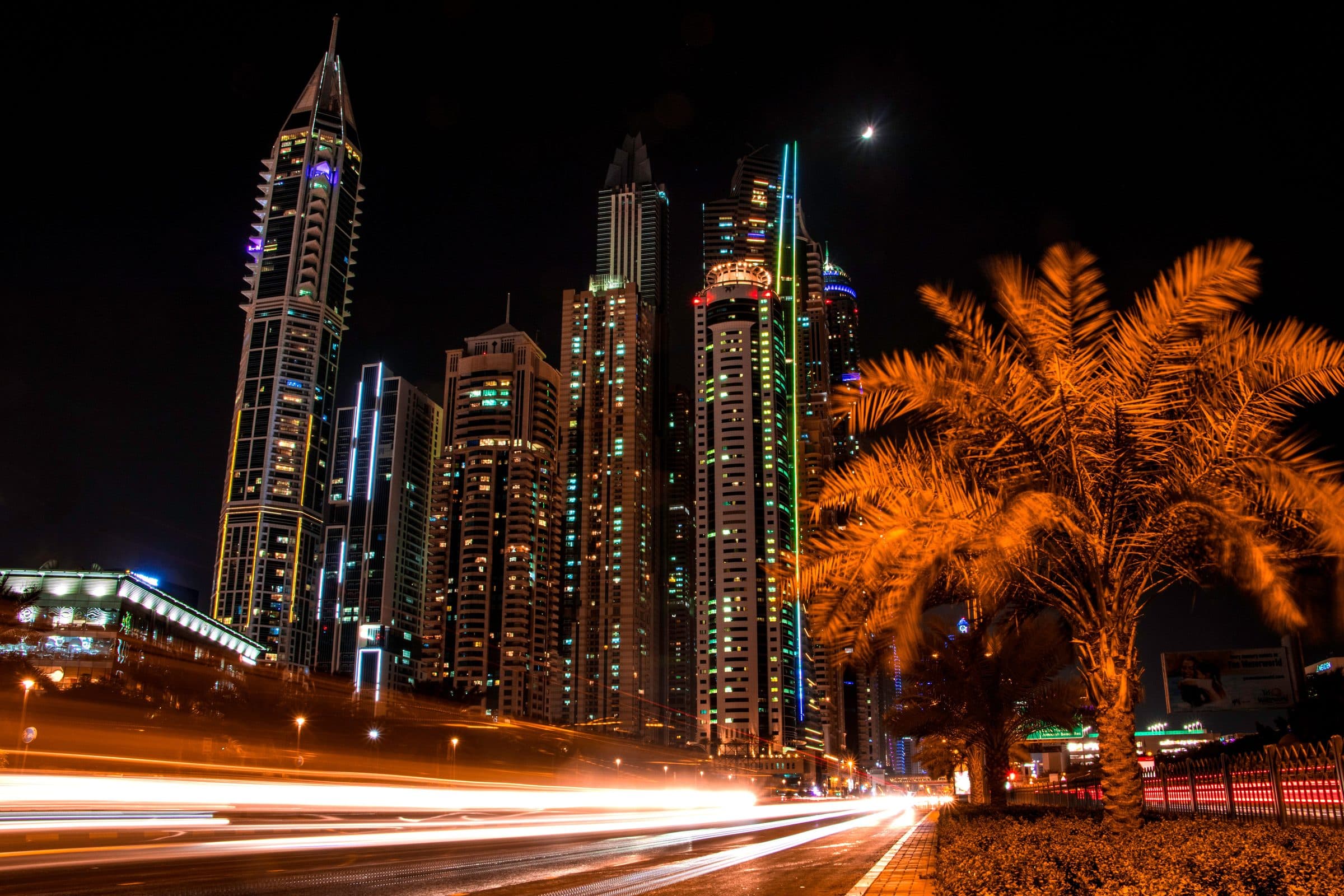 Vista noturna de Dubai com arranha-céus iluminados, palmeiras e rastros de luz de veículos
