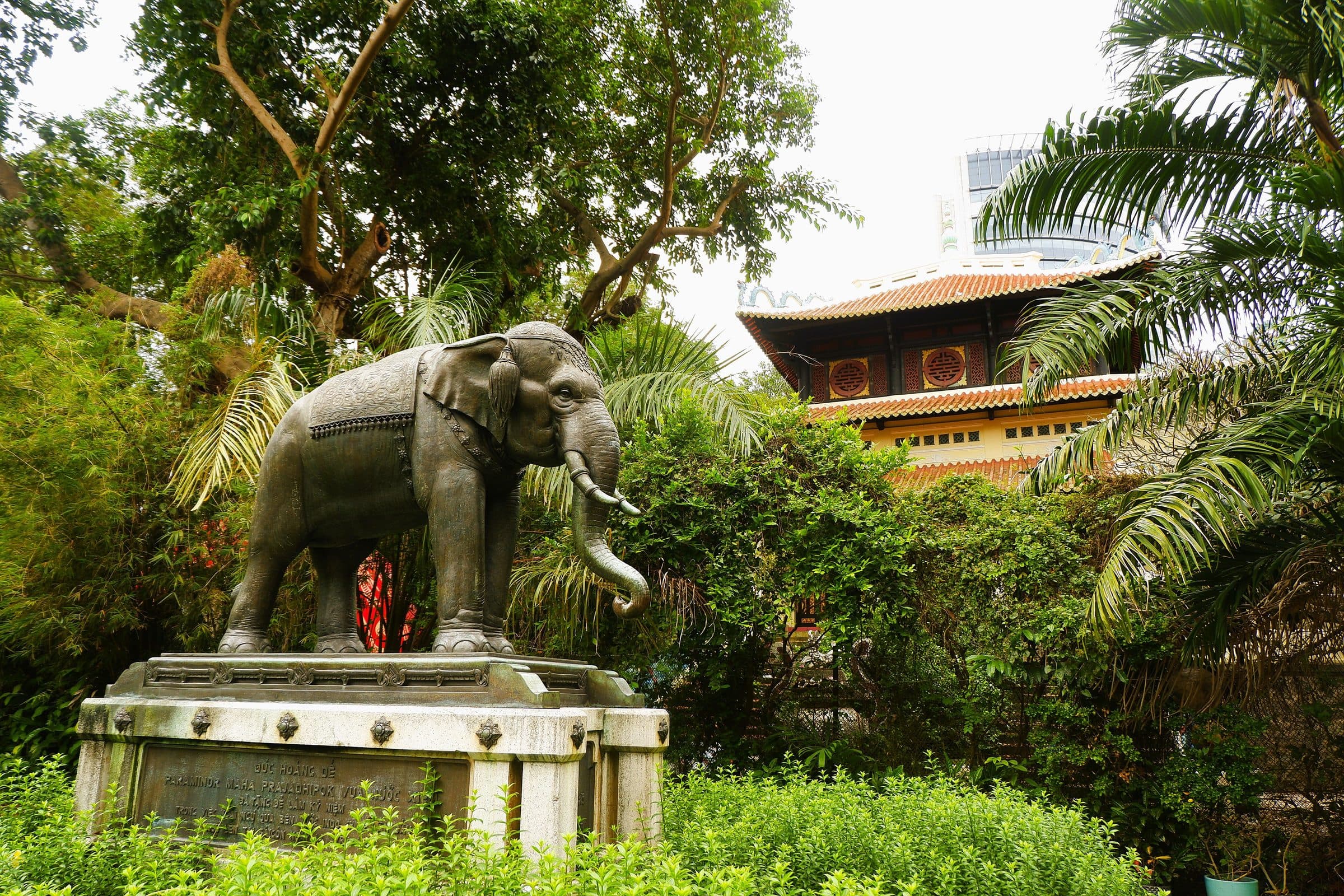 Estátua de elefante de bronze cercada por vegetação e arquitetura tradicional no Saigon Zoo