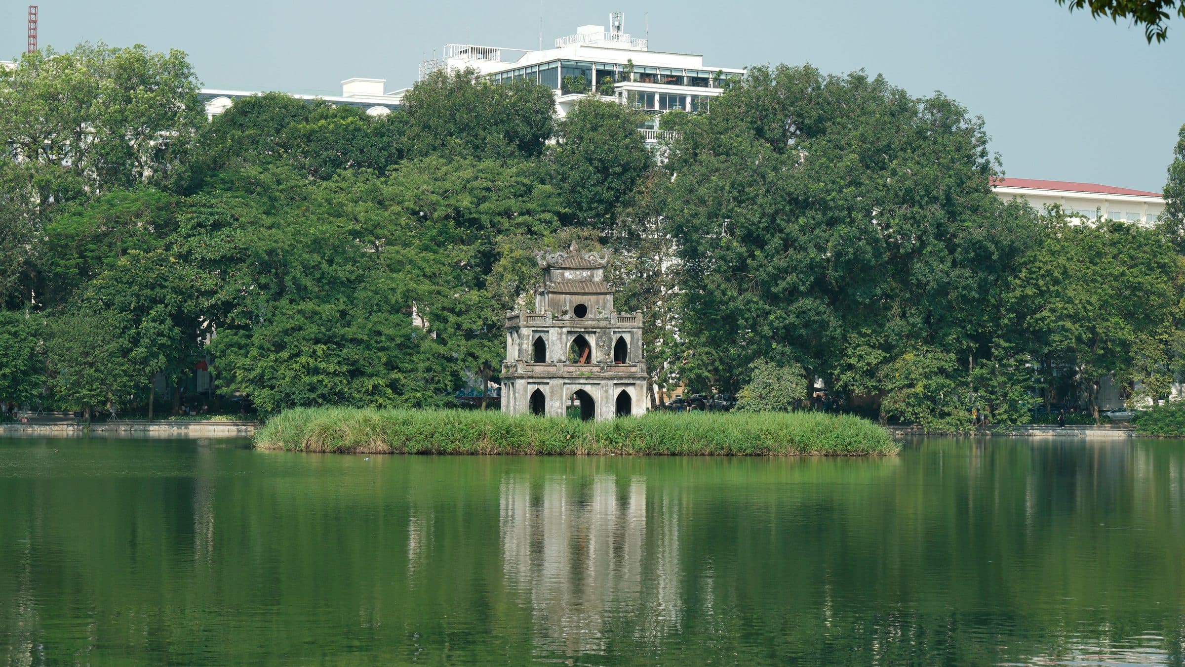Turtle Tower refletida nas águas do Lago Hoan Kiem cercada por árvores em dia ensolarado