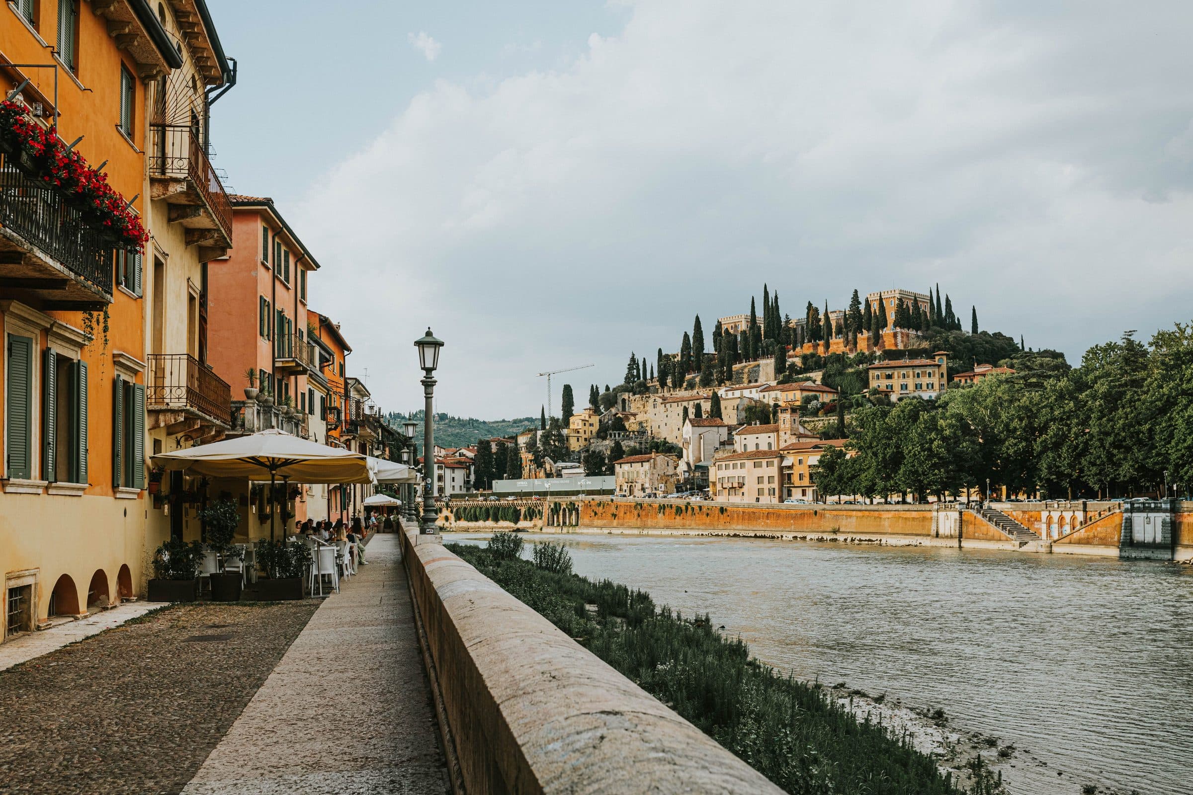Calçadão à beira do rio em Verona com prédios coloridos e vegetação