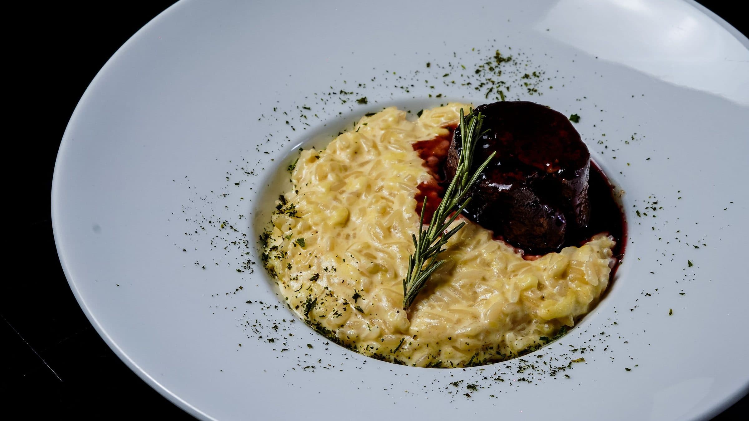 Prato branco servido com risoto, pedaços de carne e um ramo de alecrim