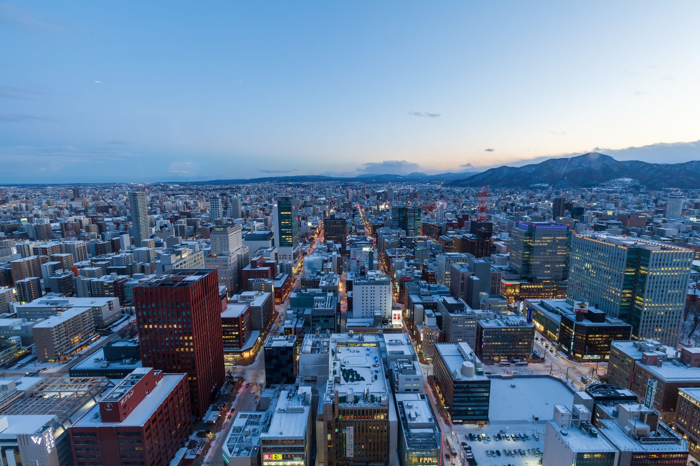 Vista aérea de Sapporo no inverno ao entardecer com prédios cobertos de neve e montanhas ao fundo