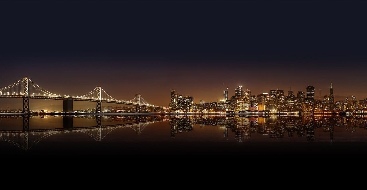 Vista panorâmica noturna de San Francisco com ponte pênsil e prédios iluminados refletidos na água