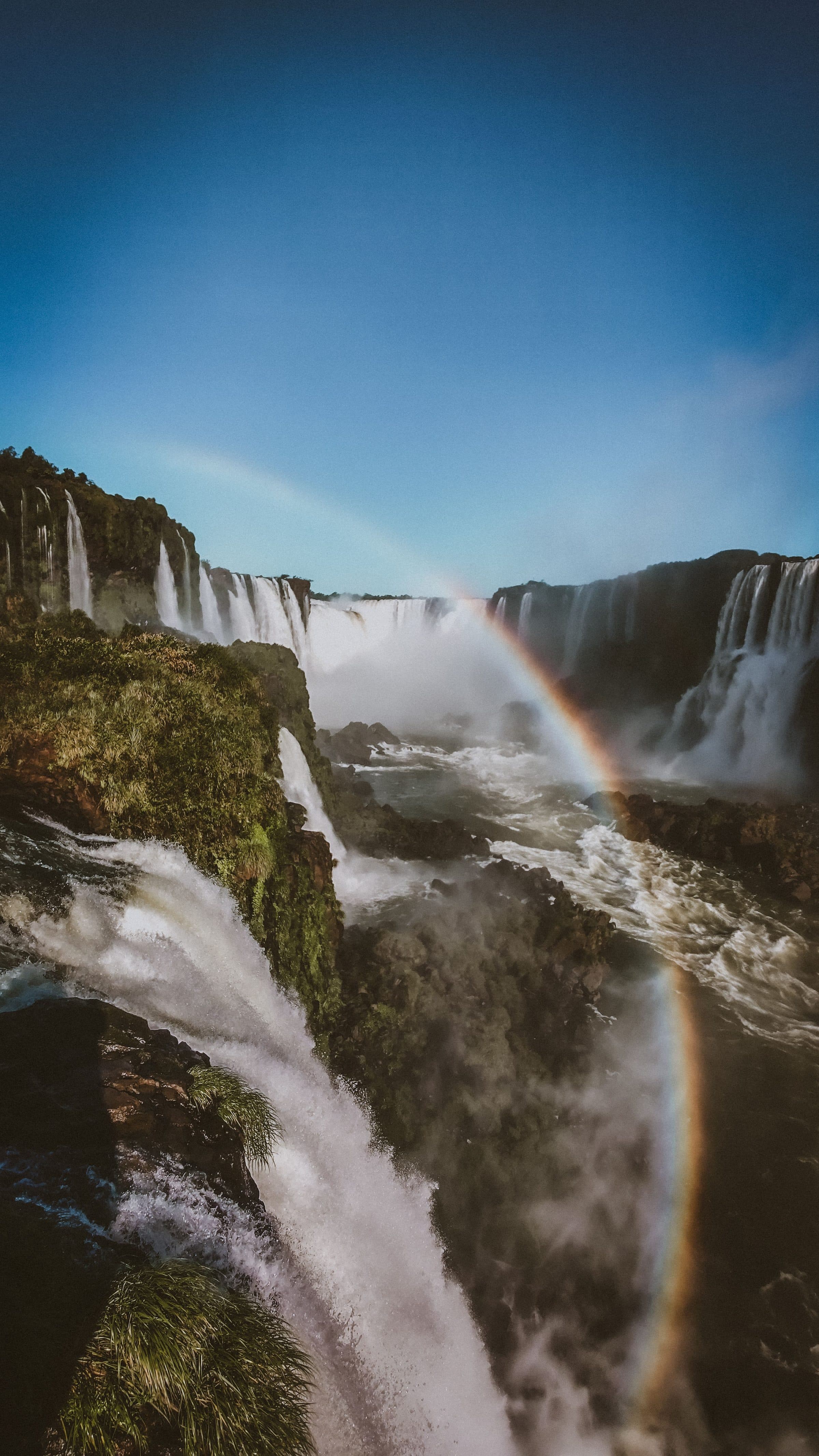 Vista das Cataratas do Iguaçu com um arco-íris sob o céu azul