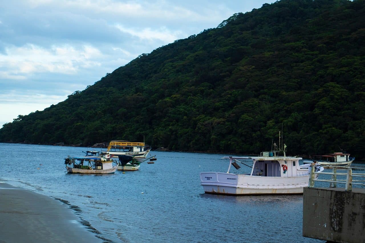 Praia em Bertioga com barcos no mar, faixa de areia e montanhas ao fundo
