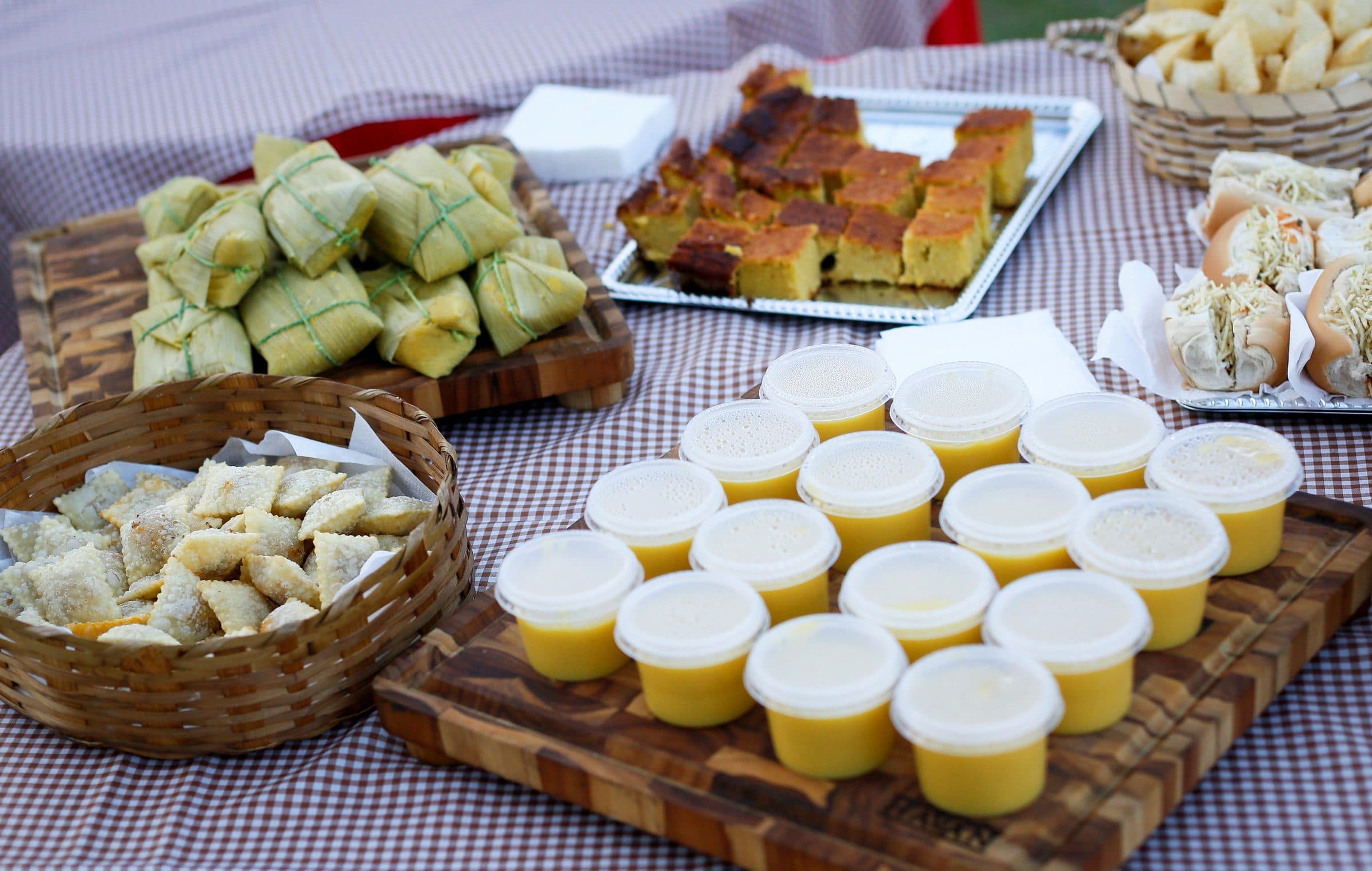 Mesa com toalha xadrez servida com bolo de milho, pamonhas e sobremesas em copinhos