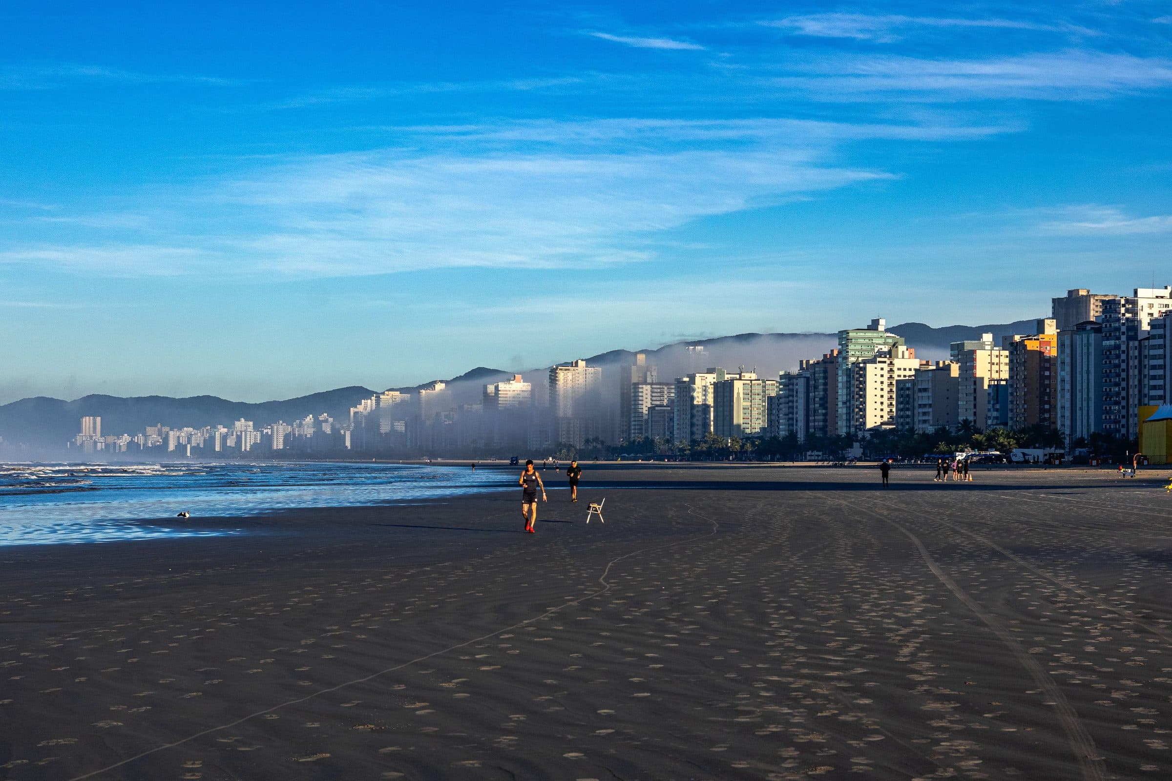 Orla de Praia Grande pela manhã com pessoas correndo e edifícios altos sob céu azul