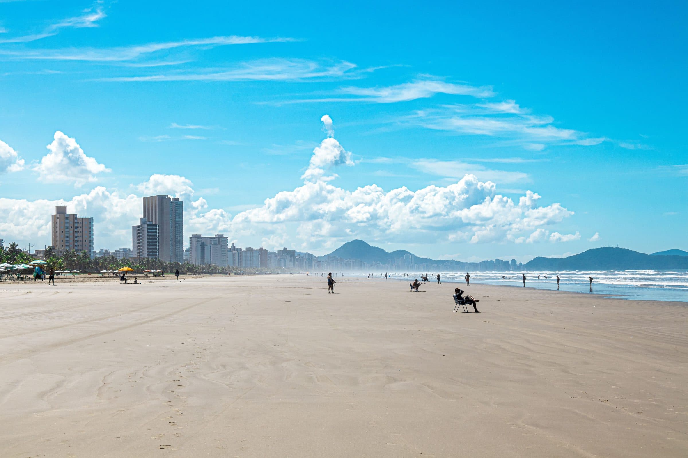 Praia urbana de Praia Grande em dia ensolarado, com prédios da cidade e montanhas ao fundo